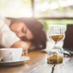 woman sleeping at desk with coffee and hourglass