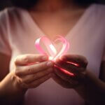 A close - up photography focusing on a woman's hands forming a heart shape with a pink ribbon against a soft pink background. scout breast cancer treatment