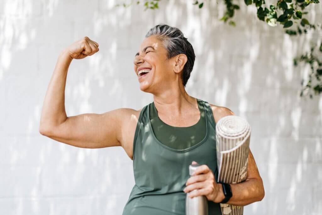 Happy elderly woman celebrating her fitness achievement after a great outdoor workout session, she flaunts her strong bicep while holding her exercise mat. Fit senior woman expressing her pride in her successful exercise routine.