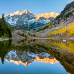 Sunrise over the iconic Maroon Bells mountains near Aspen, Colorado. Clear sky above with colorful aspen trees in full fall foliage color splendor with the entire scene reflected in the small lake in the foreground.