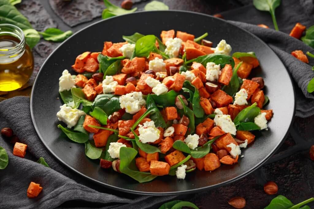 Healthy roasted sweet potato salad with spinach, feta cheese, hazelnut nuts in black plate, rustic background.